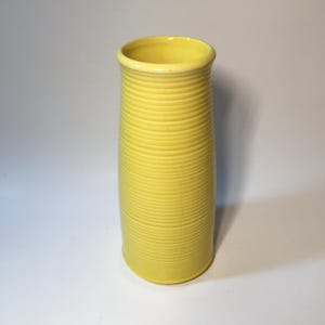 May include: A tall, cylindrical ceramic vase in a bright yellow hue. The exterior surface features a continuous, fine horizontal ribbed texture that runs from the base to the slightly flared rim, creating a uniform, ridged appearance.