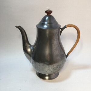 May include: A vintage metal teapot with a curved spout and a woven handle. The teapot has a dark, aged appearance with a wooden knob on the lid. The handle is made of woven light brown material.
