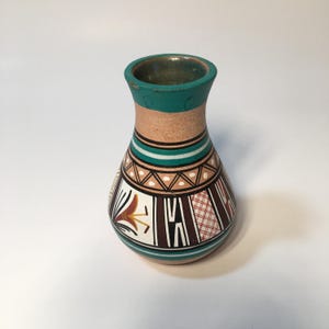 May include: A small ceramic vase with a flared rim, featuring a teal neck and base. The body is decorated with geometric patterns in black, white, and brown, including a stylized floral motif, a cross-hatched section, and a band of triangles.