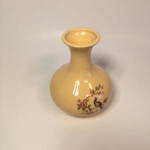 May include: A pale yellow ceramic vase with a round body and a narrow neck. The vase has a floral design with a bird on a branch.