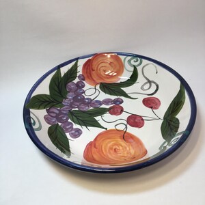 May include: A white ceramic serving bowl with a blue rim, decorated with hand-painted fruit and leaves. The bowl features grapes, cherries, and oranges. The fruit is painted in shades of purple, red, and orange, with green leaves.