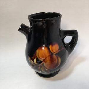 May include: Black ceramic pitcher with a spout and handle. The pitcher features a painted design of brown and orange fruit with green leaves. The pitcher has a glossy finish and sits on a small, round base. The overall shape is rounded with a wide opening.