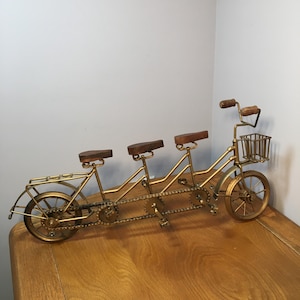 May include: A gold-colored metal tandem bicycle sculpture with three seats and a front basket. The seats and handlebars are made of wood. The bicycle has a chain and gears, and is displayed on a wooden surface.