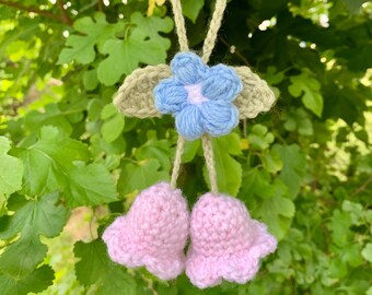 May include: A crocheted hanging ornament with a blue flower and two pink bells. The ornament is made with yarn and has a light brown string.