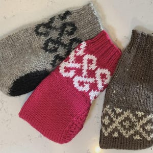 Op de afbeelding: Drie gebreide hondentruien in grijs, roze en bruin. De grijze trui heeft een zwart accent en een zwart-grijs patroon. De roze trui heeft een wit patroon. De bruine trui heeft een wit en bruin patroon.