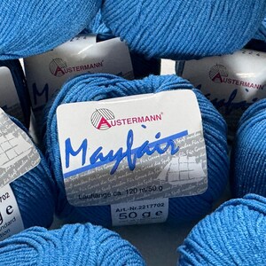 Puede incluir: Primer plano de varios ovillos de hilo azul etiquetados como "Mayfair" por Austermann. La etiqueta indica que el hilo tiene 120 metros por 50 gramos.