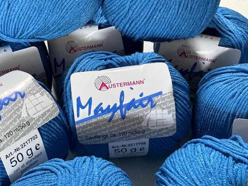 Puede incluir: Primer plano de varios ovillos de hilo azul etiquetados como "Mayfair" por Austermann. La etiqueta indica que el hilo tiene 120 metros por 50 gramos.