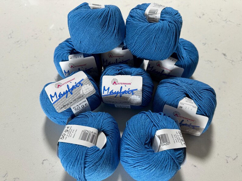 Puede incluir: Una colecci&oacute;n de diez ovillos de lana azul con etiquetas blancas que dicen "Mayfair" y "Austermann". Cada ovillo pesa 50 gramos.
