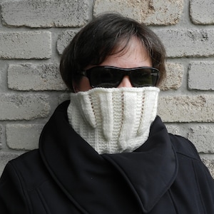 May include: A person wearing a black coat with a white knitted neck warmer and black sunglasses.
