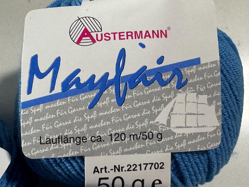 Puede incluir: Una madeja de lana azul con la etiqueta "Mayfair" y la marca "USTERMANN". El texto "Laufl&auml;nge ca. 120 m/50 g" y "Art.-Nr. 2217702 50 ge" tambi&eacute;n es visible.