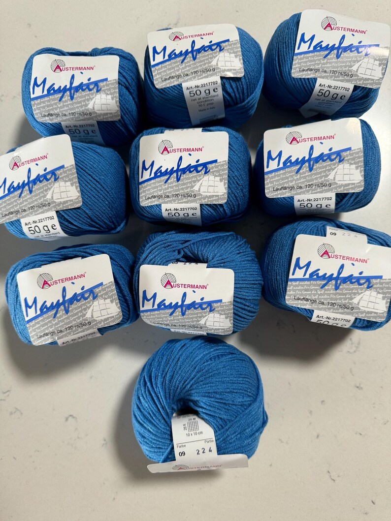 Puede incluir: Diez ovillos de hilo azul etiquetados como "Mayfair" por Austermann. Cada ovillo pesa 50 gramos y tiene una longitud de 120 metros. El hilo es de peso medio y es adecuado para tejer o hacer ganchillo.