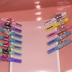 May include: A collection of colorful hair clips with a crown design and the word "QUEEN" printed on them. The clips are in various colors, including pink, purple, blue, and yellow, with silver and rhinestone accents.