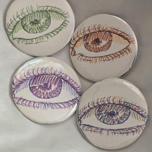 Könnte beinhalten: Vier runde, weiße Knöpfe mit handgezeichneten Augendesigns. Jedes Auge ist unterschiedlich gefärbt: grün, braun, lila und blau. Die Knöpfe sind auf einer weißen, leicht strukturierten Oberfläche angeordnet.