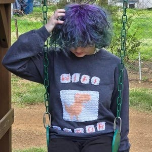 May include: A dark grey sweatshirt featuring a white and peach crochet design. The design includes a rectangular shape with a peach centre and several smaller crochet shapes above and below.