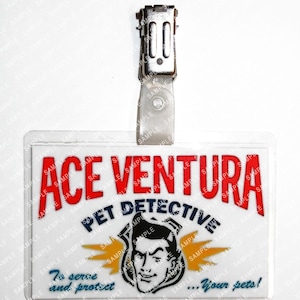 Op de afbeelding: Een doorzichtige plastic naamtag met een metalen clip. De tag bevat de tekst "ACE VENTURA PET DETECTIVE" in rood en blauw, met een cartoonafbeelding van een man en de zin "To serve and protect...your pets!"