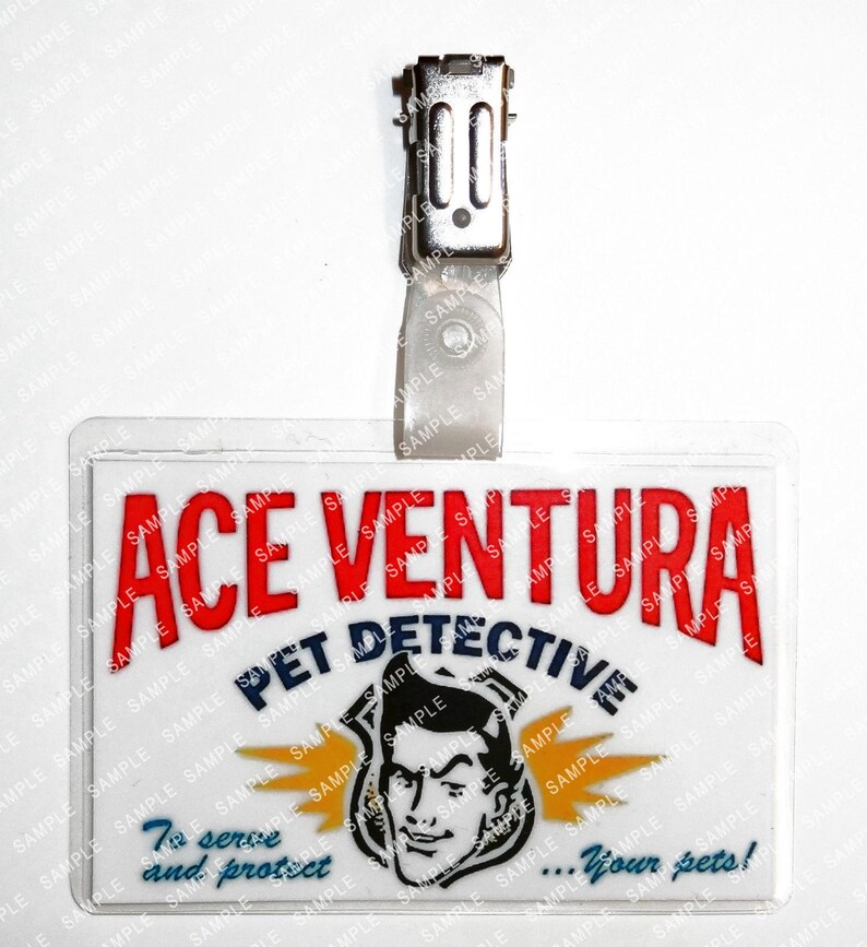 Op de afbeelding: Een doorzichtige plastic naamtag met een metalen clip. De tag bevat de tekst "ACE VENTURA PET DETECTIVE" in rood en blauw, met een cartoonafbeelding van een man en de zin "To serve and protect...your pets!"