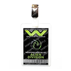 May include: A clear plastic ID badge with a metal clip. The badge has a black background with green text that reads "WEYLAND-YUTANI CORP" and "WEAPONS SPECIALIST ALIEN DIVISION". The badge also features a barcode and the text "BUILDING BETTER WORLDS".