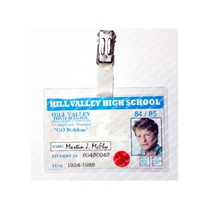 Puede incluir: Una tarjeta de identificación de plástico blanco con un borde azul y el texto "Hill Valley High School" impreso en ella. La tarjeta tiene una foto de un joven con cabello castaño y un círculo rojo con el texto "84/85" impreso en ella. La tarjeta también tiene el texto "Martin J. McFly" impreso en ella, junto con un número de identificación de estudiante y el año "1984-1985".