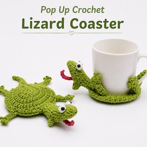 Op de afbeelding: Twee groene gehaakte hagedis-onderzetters met wit-zwarte ogen en roze tongen. De ene ligt plat, terwijl de andere om een witte keramische mok heen zit. De tekst Pop Up Crochet Lizard Coaster staat bovenaan de afbeelding.