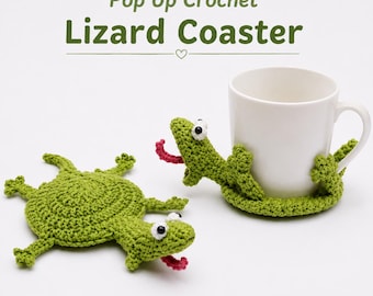 May include: Two green crochet lizard coasters with white and black eyes and pink tongues. One coaster lies flat, while the other wraps around a white ceramic mug. The text "Pop Up Crochet Lizard Coaster" is displayed above the items.