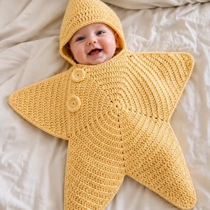 Op de afbeelding: Een felgele gehaakte babyslaapzak in de vorm van een vijfpuntige ster. Het kledingstuk is voorzien van een puntige capuchon, een gestructureerd steekpatroon dat vanuit het midden uitstraalt en twee ronde knopen aan de linkerbovenkant.