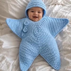 May include: A light blue crocheted baby bunting shaped like a five-pointed star. The garment features a pointed hood and two circular buttons on the upper right side. The texture is consistent throughout, showcasing a detailed yarn stitch pattern.