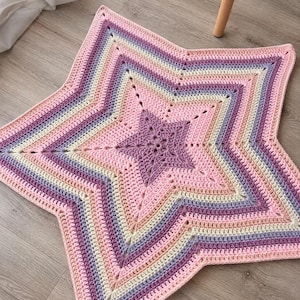 Può includere: Un tappeto all'uncinetto a forma di stella con bande concentriche di colore nelle tonalità viola, rosa chiaro e crema. Il centro è una stella viola tinta unita, circondata da file alternate di punti che si espandono verso l'esterno.