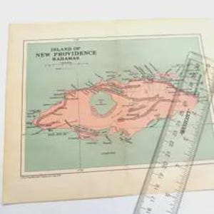 May include: A vintage map titled Island of New Providence Bahamas with a salmon-coloured landmass on a light green background, featuring black text and lines. A clear plastic ruler is placed diagonally across the map, showing a length of 25.4 cm.