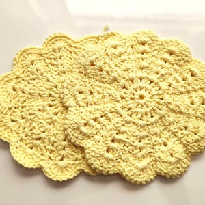 May include: Two round, pale yellow crochet doilies featuring a detailed, intricate floral-inspired pattern with a scalloped edge. The circular mats are crafted with a textured, knotted stitch design, creating a layered, decorative appearance.
