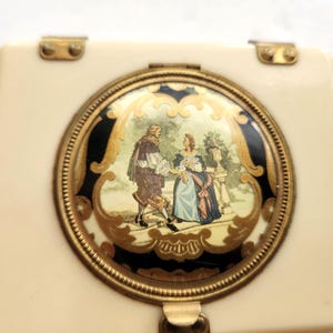 May include: Ivory-coloured vintage compact with a gold-toned frame and clasp. The compact features a circular design with a painted scene of figures in period clothing. The frame has a black and gold decorative border.