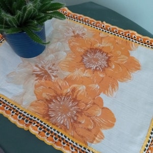 May include: A white crocheted table runner with a yellow and orange floral design. The runner has a crocheted border with a scalloped edge.