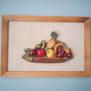 May include: A framed relief sculpture of a fruit basket. The basket contains a pineapple, bananas, apples, grapes, and other fruits. The frame is made of light brown wood, and the background is a light beige fabric.