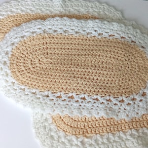 May include: Two oval-shaped crochet doilies featuring a central section of light peach yarn surrounded by a wide, textured border of white yarn. The crochet work displays a consistent stitch pattern throughout, creating a layered, decorative appearance.