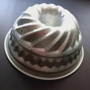 Puede incluir: Un molde para bizcocho Bundt de color plateado con un diseño decorativo acanalado. El molde tiene forma circular con un agujero central y un borde festoneado. El molde es de metal y está diseñado para hornear pasteles.