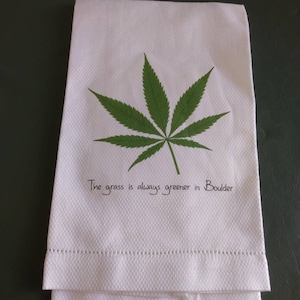 May include: White kitchen towel with a green marijuana leaf printed on it. The text "The grass is always greener in Boulder" is printed below the leaf.
