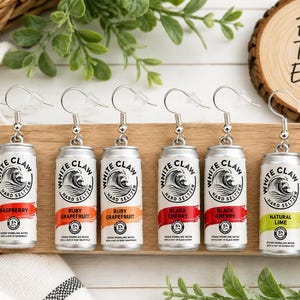 May include: Eight miniature White Claw hard seltzer can earrings with silver-toned hooks. The pairs feature Raspberry, Ruby Grapefruit, Black Cherry, and Natural Lime flavors, each displaying the brand logo, flavor name, and 5% alcohol content text.