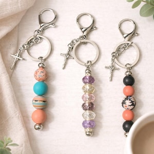 May include: Three handmade keychains with silver-tone hardware and beaded accents. Each keychain features a cross charm and a series of colorful beads. The beads include shades of pink, blue, orange, and black, with patterned and solid designs.