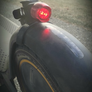 May include: Close-up of a black electric scooter with a red tail light. The scooter has a black tire with a yellow stripe. The text "Tail light not included" is visible in the image.