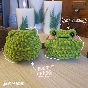 May include: Two handmade green crochet frog plushies. One frog has a round shape, and the other has a face with black eyes and pink cheeks. The text "BOOTYlicious" and "BOOTY = FROG" are written on the image.