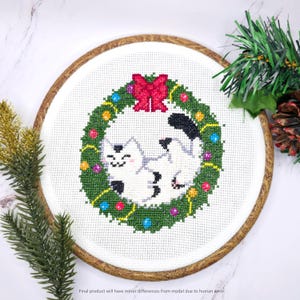 May include: Cross-stitch embroidery of a white cat with black spots, inside a green wreath with a red bow. The wreath is decorated with colorful ornaments. The embroidery is set in a wooden hoop, with text at the bottom: "Final product will have minor differences from model due to human error."