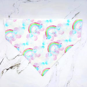 May include: White triangular pet bandana with a watercolor rainbow and cloud pattern. The rainbows have red, orange, yellow, green, and blue arches. The clouds are pink and blue. The bandana is made of a soft fabric.