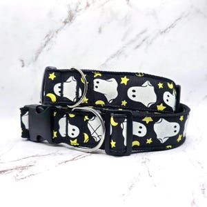 May include: Two black dog collars with a white ghost, yellow star, and moon pattern. Each collar has a black plastic buckle and a silver metal D-ring for leash attachment. The collars are stacked on top of each other.