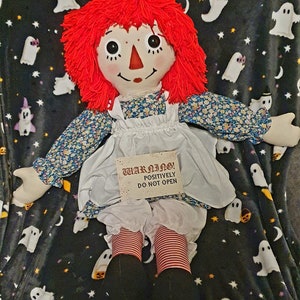A rag doll with red yarn hair, a painted face, and a floral print dress. The doll holds a sign saying "WARNING! POSITIVELY DO NOT OPEN." It is set against a black blanket with white ghost and star patterns.
