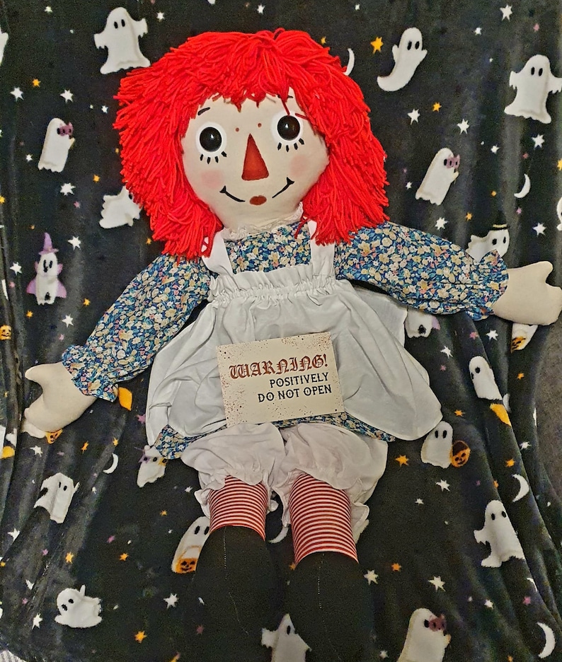 A rag doll with red yarn hair, a painted face, and a floral print dress. The doll holds a sign saying "WARNING! POSITIVELY DO NOT OPEN." It is set against a black blanket with white ghost and star patterns.