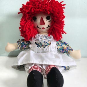 Handmade rag doll with bright red yarn hair, embroidered facial features, floral cotton dress, white apron, striped socks and black shoes, seated for display.