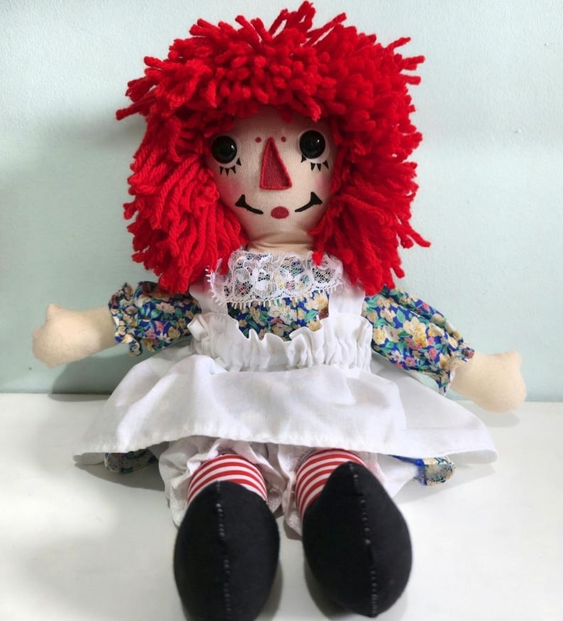 Handmade rag doll with bright red yarn hair, embroidered facial features, floral cotton dress, white apron, striped socks and black shoes, seated for display.