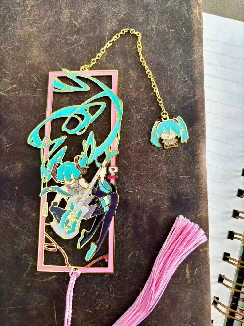 K&ouml;nnte beinhalten: Ein pinkfarbener und goldfarbener Metall-Lesezeichen mit einem t&uuml;rkisfarbenen Anime-Charakter, der Gitarre spielt. Das Lesezeichen hat einen passenden Anh&auml;nger an einer goldenen Kette. Am unteren Rand des Lesezeichens ist eine pinkfarbene Quaste befestigt.