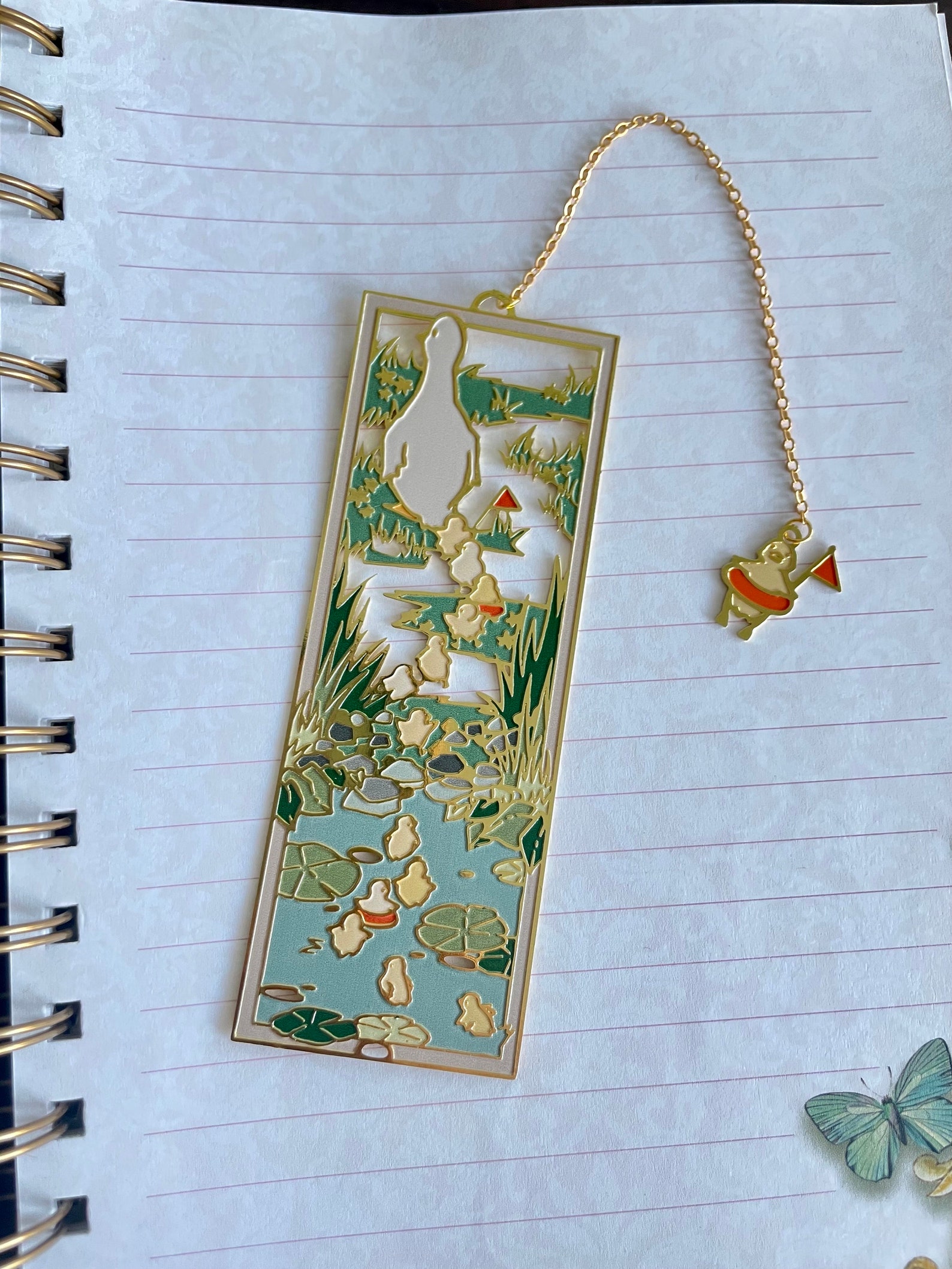 An ornate enamel bookmark with a trail of ducklings following a duck across a pond. It is sitting on a piece of lined paper and has a chain with a duck on the end attached to the top.