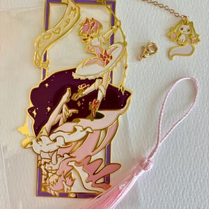 K&ouml;nnte beinhalten: Ein goldener Metall-Lesezeichen mit einer Comicfigur mit rosa Haaren und einem rosa-wei&szlig;en Kleid. Das Lesezeichen ist an einem rosa Quasten und einer goldenen Kette mit einem kleinen Anh&auml;nger befestigt.