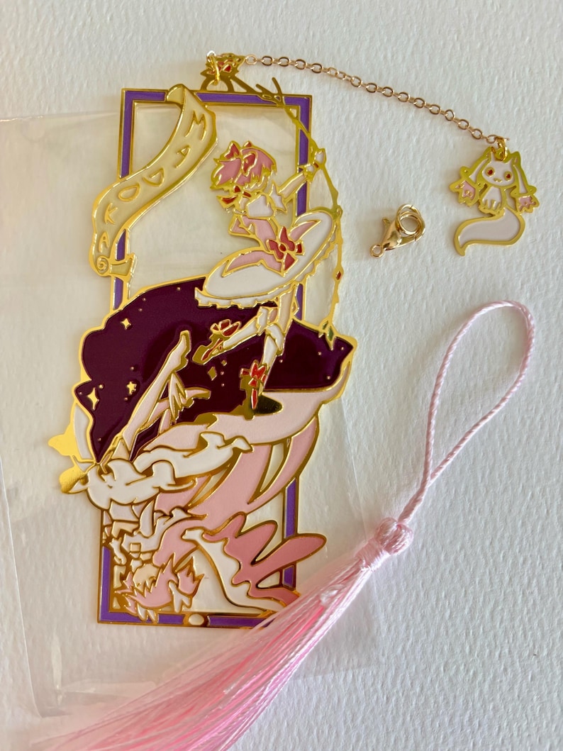 K&ouml;nnte beinhalten: Ein goldener Metall-Lesezeichen mit einer Comicfigur mit rosa Haaren und einem rosa-wei&szlig;en Kleid. Das Lesezeichen ist an einem rosa Quasten und einer goldenen Kette mit einem kleinen Anh&auml;nger befestigt.